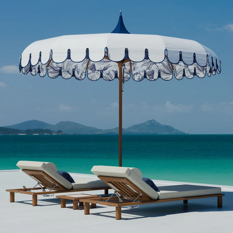 Tuuci Ocean Master M1 Voilare Wonder center post parasol with a navy-trimmed scalloped canopy and patterned lining over two sun loungers, with a turquoise sea and green islands behind