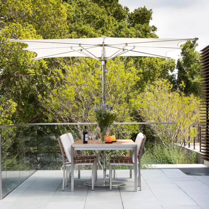 White Tuuci Ocean Master M1 Zero Horizon center post parasol with a flat canopy over a dining table and chairs on a paved terrace with a glass balustrade, surrounded by trees