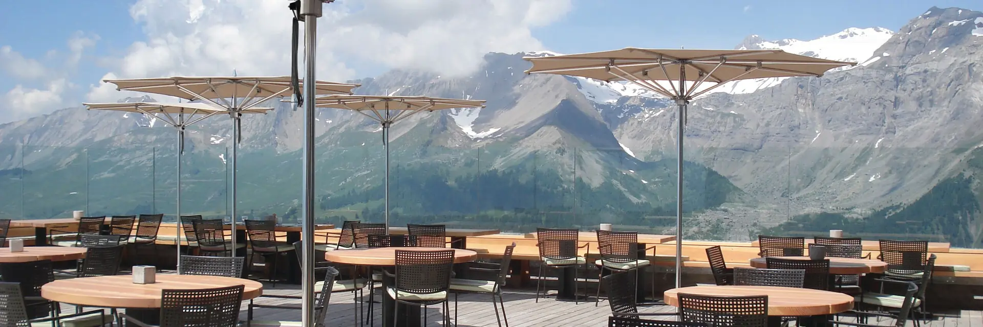 Tuuci Ocean Master M1 Zero Horizon parasols over round tables on an exposed restaurant terrace, with snow-capped mountains behind