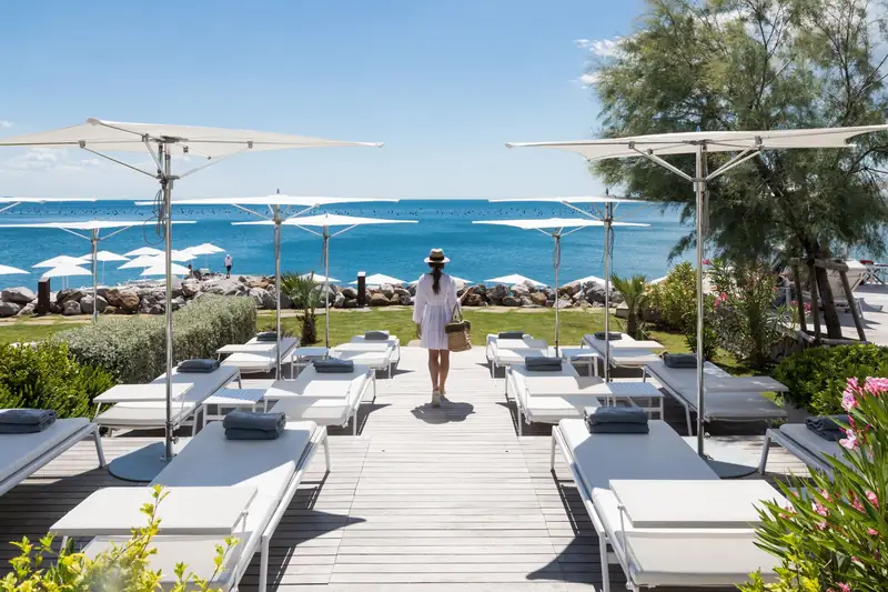 White Tuuci Ocean Master M1 Zero Horizon parasols with polished masts over white sunbeds on a seaside deck, with a woman walking toward the sea