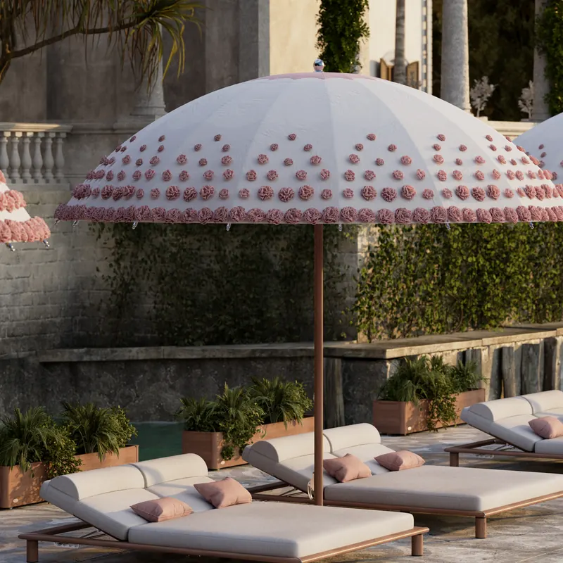 Tuuci Ocean Master MAX Bolero: Ombré Blossom center post parasol, its domed canopy covered in pink tufted blossoms, shading cushioned sun loungers on a stone terrace with ivy-covered walls