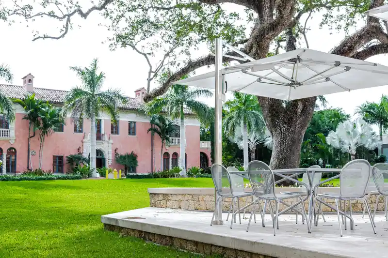 White Tuuci Ocean Master MAX single cantilever parasol over a table and chairs on a garden terrace, with a pink villa behind