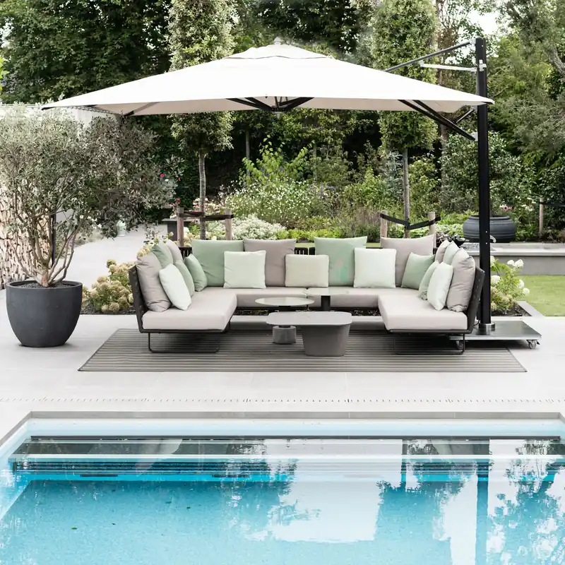 Off-white Tuuci Ocean Master MAX Classic Cantilever parasol on a black side mast shading a U-shaped sectional sofa beside a swimming pool in a landscaped garden
