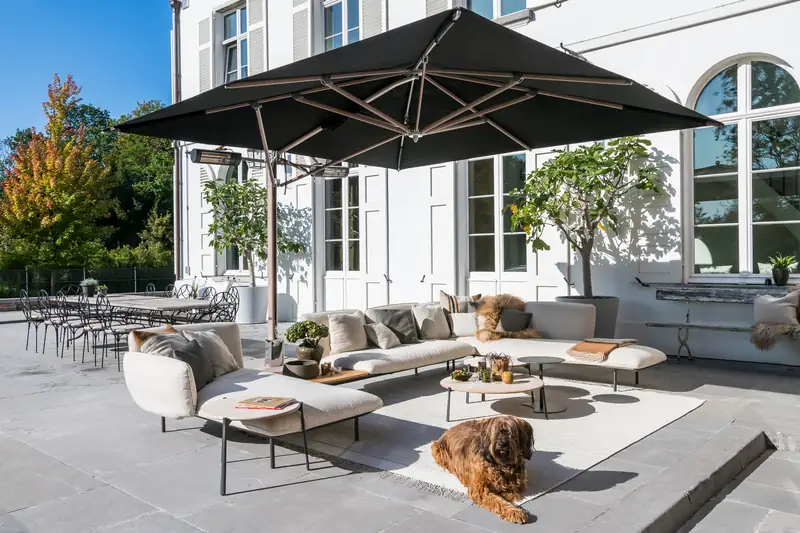 Black Tuuci Ocean Master MAX Classic Cantilever parasol with heaters under its canopy, shading a curved lounge sofa on the stone terrace of a white villa