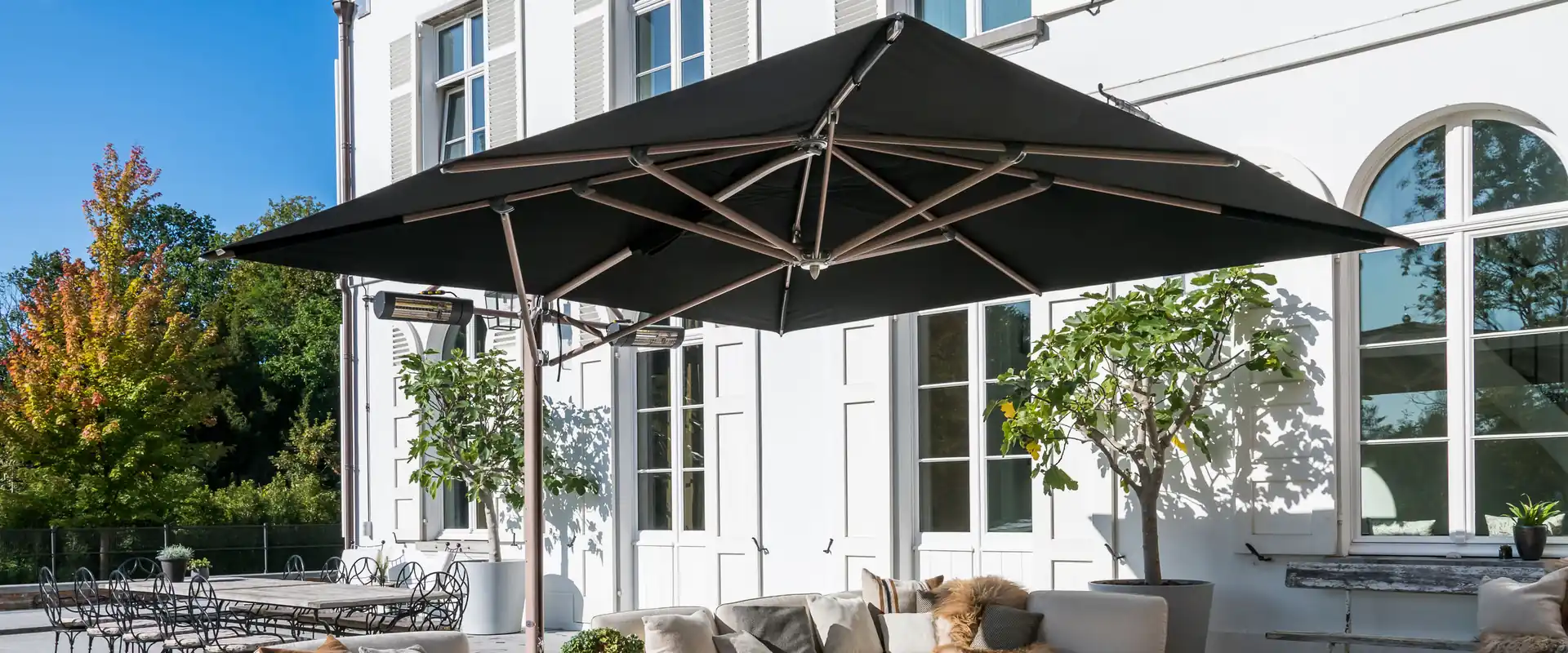 Black Tuuci Ocean Master MAX Classic Cantilever parasol with heaters under its canopy, shading a curved lounge sofa on the stone terrace of a white villa