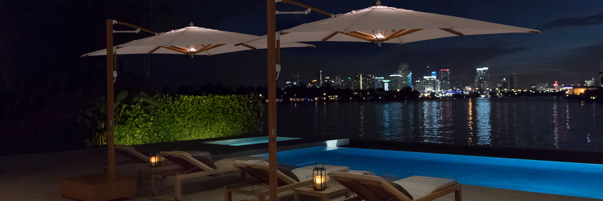 Two Tuuci Ocean Master MAX cantilever parasols with built-in lights over sun loungers beside a pool at night, with a city skyline across the water