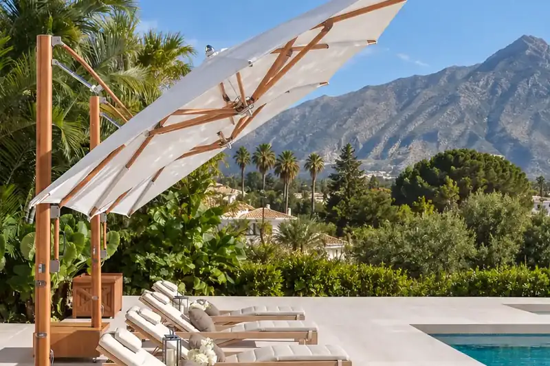 Two white Tuuci Ocean Master MAX Classic Cantilever parasols with teak-finish masts over sun loungers beside a villa pool, with La Concha mountain behind
