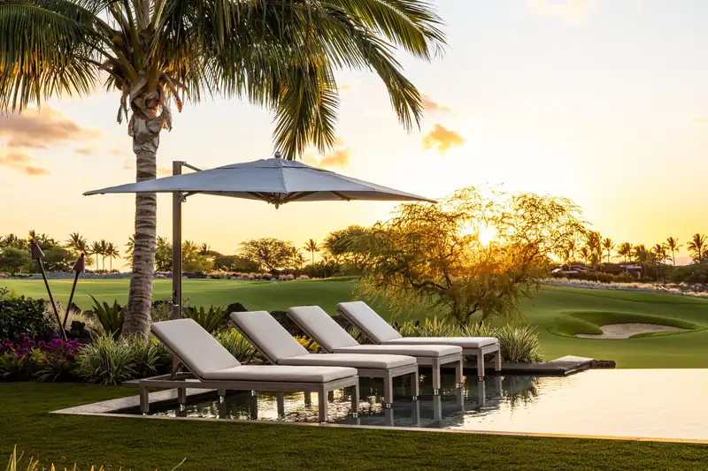 Tuuci Ocean Master MAX Classic Cantilever parasol shading a row of cushioned sun loungers, with a palm tree and a golf course behind at sunset