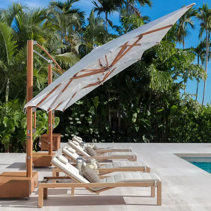 White Tuuci Ocean Master MAX Classic Cantilever parasol with a wood-finish side mast, tilted to shade a row of sun loungers on a pale stone pool terrace backed by palms