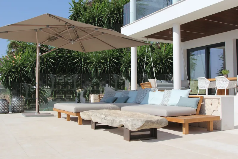 Taupe Tuuci Ocean Master MAX Classic Cantilever parasol shading a teak sofa with pale blue cushions on the terrace of a white villa, with palms behind