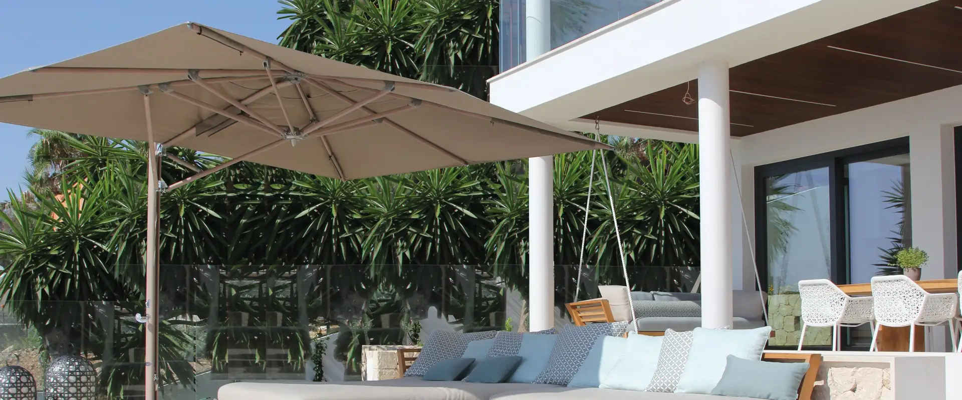 Taupe Tuuci Ocean Master MAX Classic Cantilever parasol shading a teak sofa with pale blue cushions on the terrace of a white villa, with palms behind