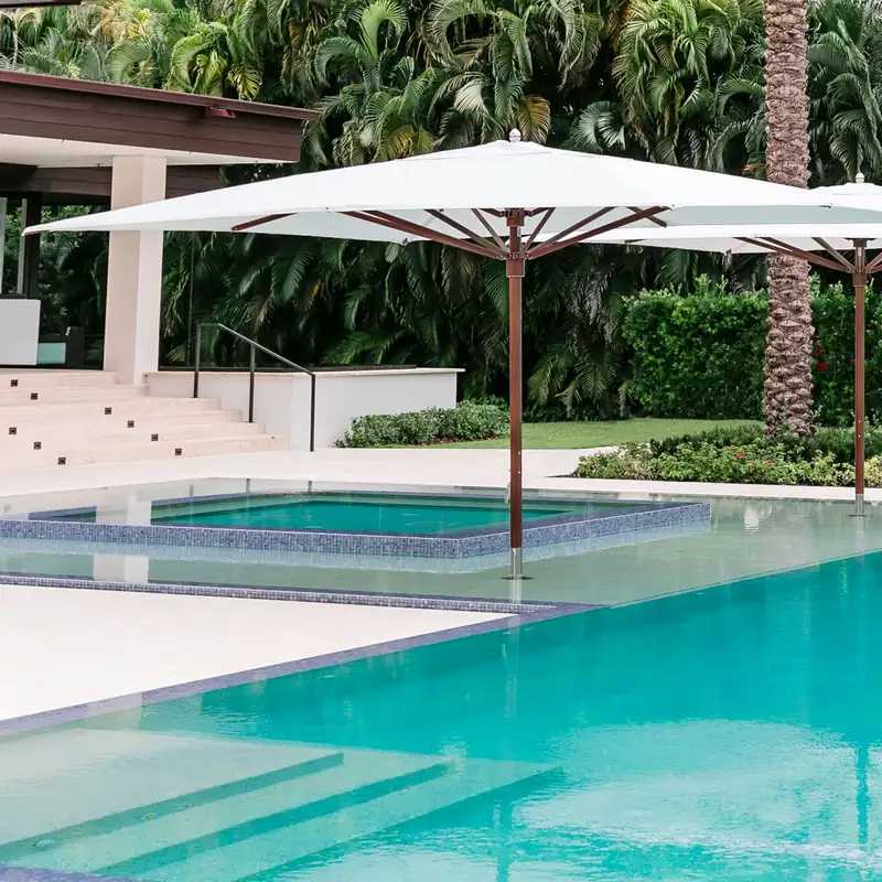 Large white square Tuuci Ocean Master MAX Classic center post parasol with a wood-finish mast standing in the shallow end of a turquoise pool, with a second one behind and dense palms beyond
