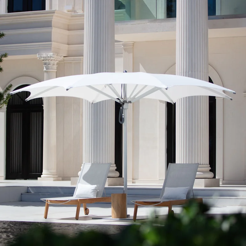 White Tuuci Ocean Master MAX Crescent center post parasol shading two teak sun loungers in front of a pale stone villa with fluted columns