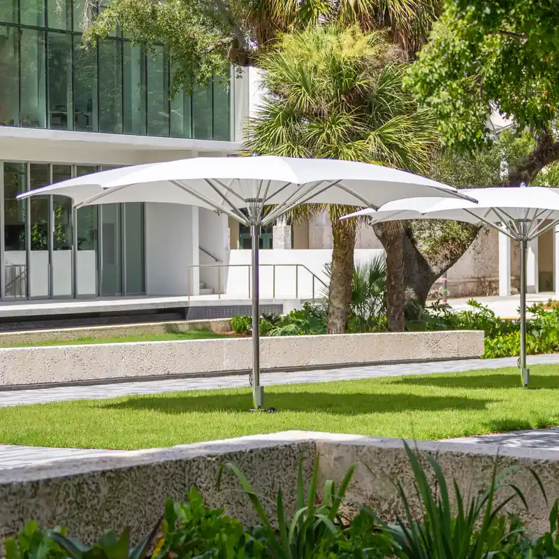 White Tuuci Ocean Master MAX Crescent center post parasols with curved canopies and polished masts on a lawn in front of a modern glass-fronted building, framed by tropical planting