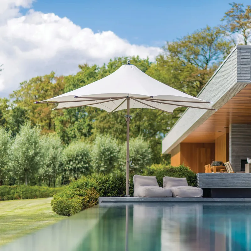 White Tuuci Ocean Master MAX F-1 center post parasol over two lounge cushions at the edge of a reflecting pool, beside a modern house clad in stone and timber, with a lawn and trees behind