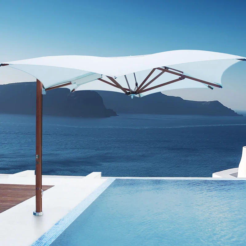 White Tuuci Ocean Master MAX Manta Cantilever parasol with a wood-finish side mast beside an infinity pool, overlooking a deep blue sea and distant cliffs