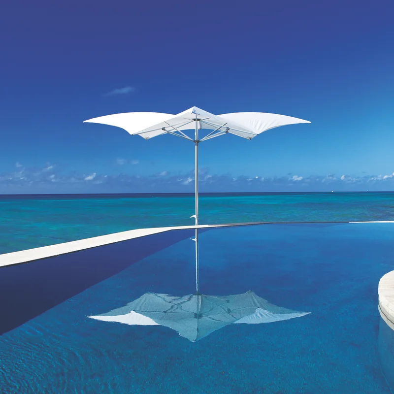 White Tuuci Ocean Master MAX Manta parasol standing at the edge of an infinity pool, reflected in the still water with the open ocean and blue sky behind