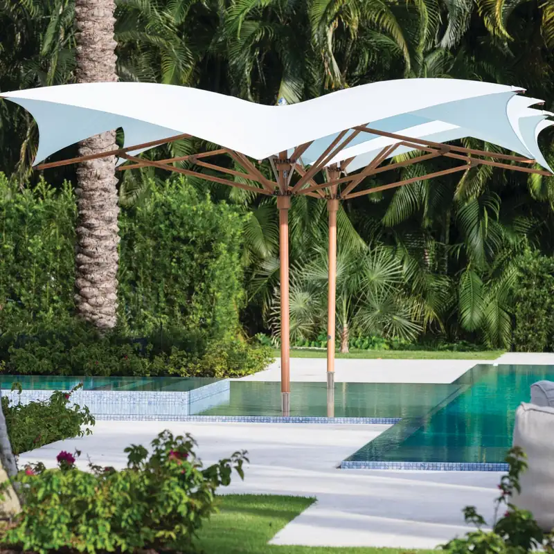 Two white Tuuci Ocean Master MAX Manta center post parasols with wood-finish masts and upswept canopy corners standing at the edge of a garden pool, surrounded by palms