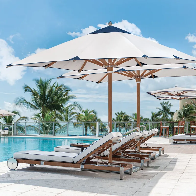 White Tuuci Ocean Master MAX Nautical Teak center post parasols with teak masts and ribs and navy trim over teak sun loungers beside a pool, with palm trees and the sea beyond