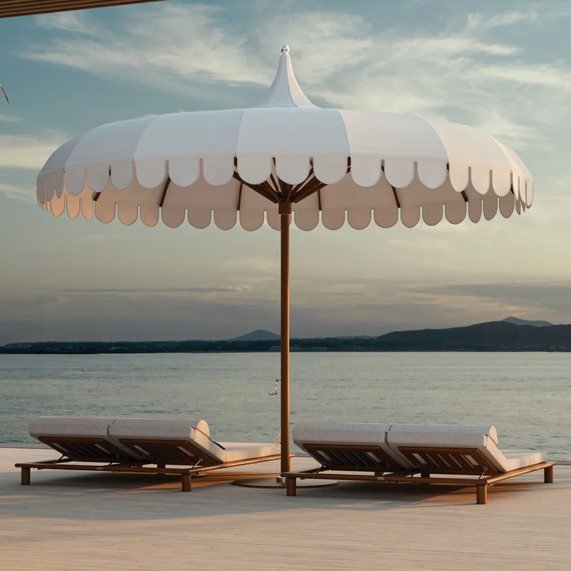 White Tuuci Ocean Master MAX Voilare center post parasol with a scalloped valance and pointed top shading two teak sun loungers on a deck by the sea at dusk