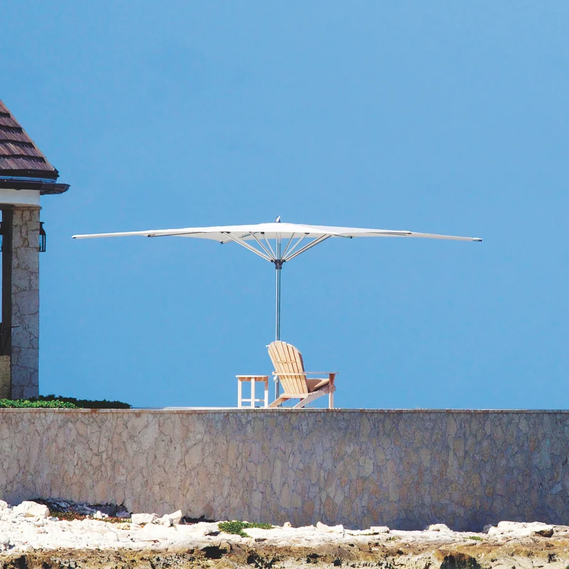 Tuuci Ocean Master MAX Zero Horizon center post parasol with a flat, level white canopy above an Adirondack chair and side table on a stone-walled terrace, against a clear blue sky