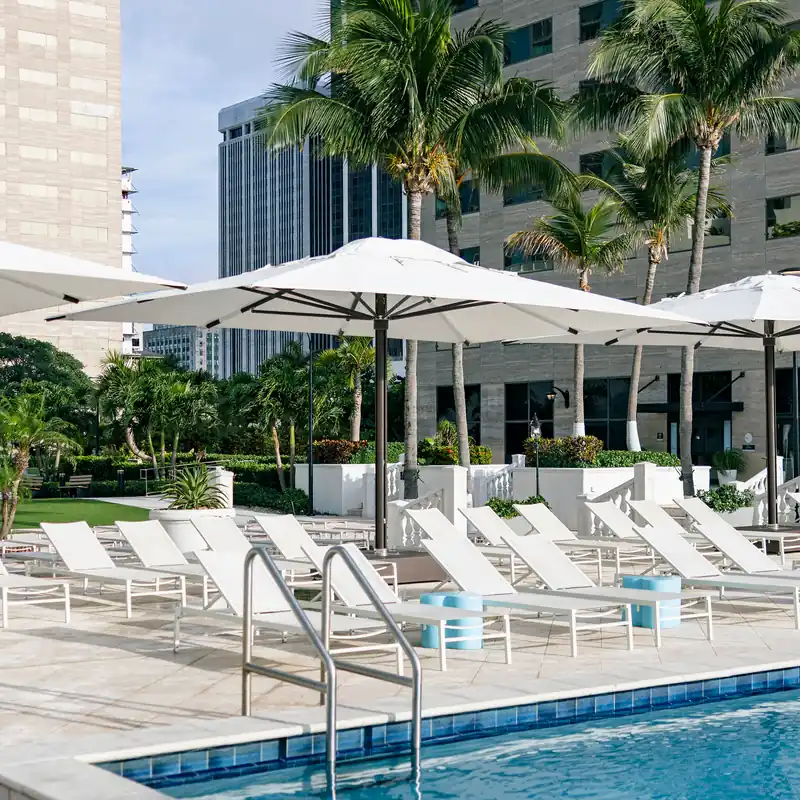 Large white rectangular Tuuci Ocean Master MEGA MAX Classic center post parasol shading a row of white sun loungers beside a hotel pool, with palm trees and high-rise buildings behind