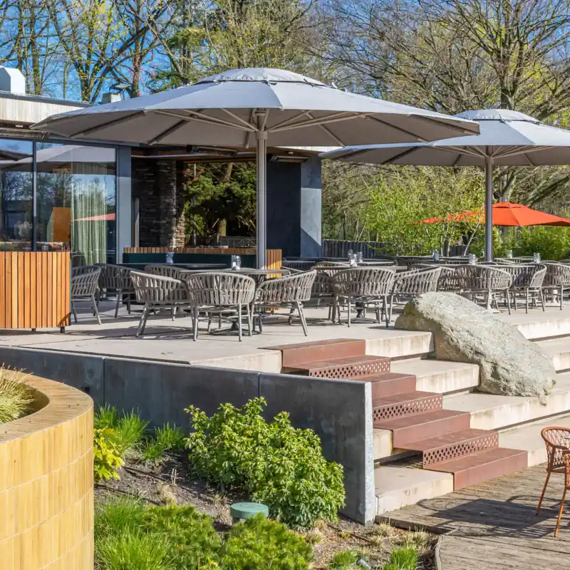 Two large round gray Tuuci Ocean Master MEGA MAX Classic center post parasols covering the tables and woven chairs of a restaurant terrace, with stone steps and trees around
