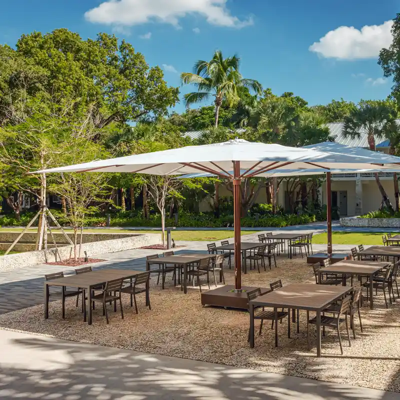 Very large white square Tuuci Ocean Master MEGA MAX Classic center post parasol with a wood-finish mast covering rows of dining tables in a garden courtyard, with a second one behind and palms beyond