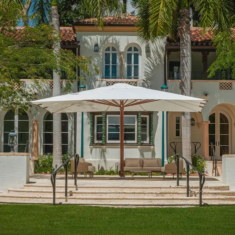Large white Tuuci Ocean Master MEGA MAX parasol covering a full seating group on the terrace of a white Mediterranean-style building with terracotta roof tiles and palm trees