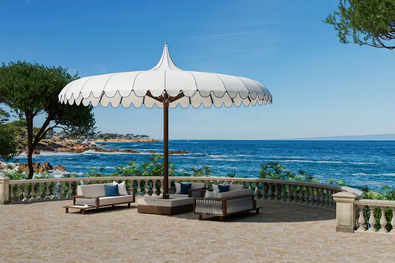 Large white Tuuci Ocean Master MEGA MAX Voilare parasol with a scalloped valance and pointed top shading a group of sofas on a stone terrace with a balustrade, overlooking the sea and pine trees