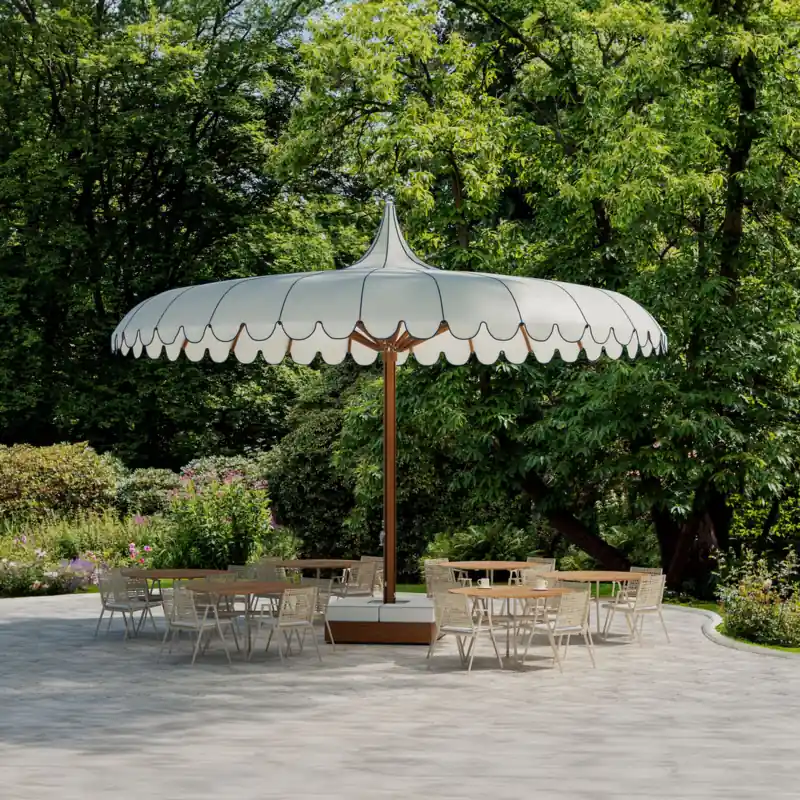 Large Tuuci Ocean Master MEGA MAX Voilare center post parasol with a scalloped white canopy, navy piping and a wood-finish mast shading bistro tables and chairs on a paved garden terrace under tall trees