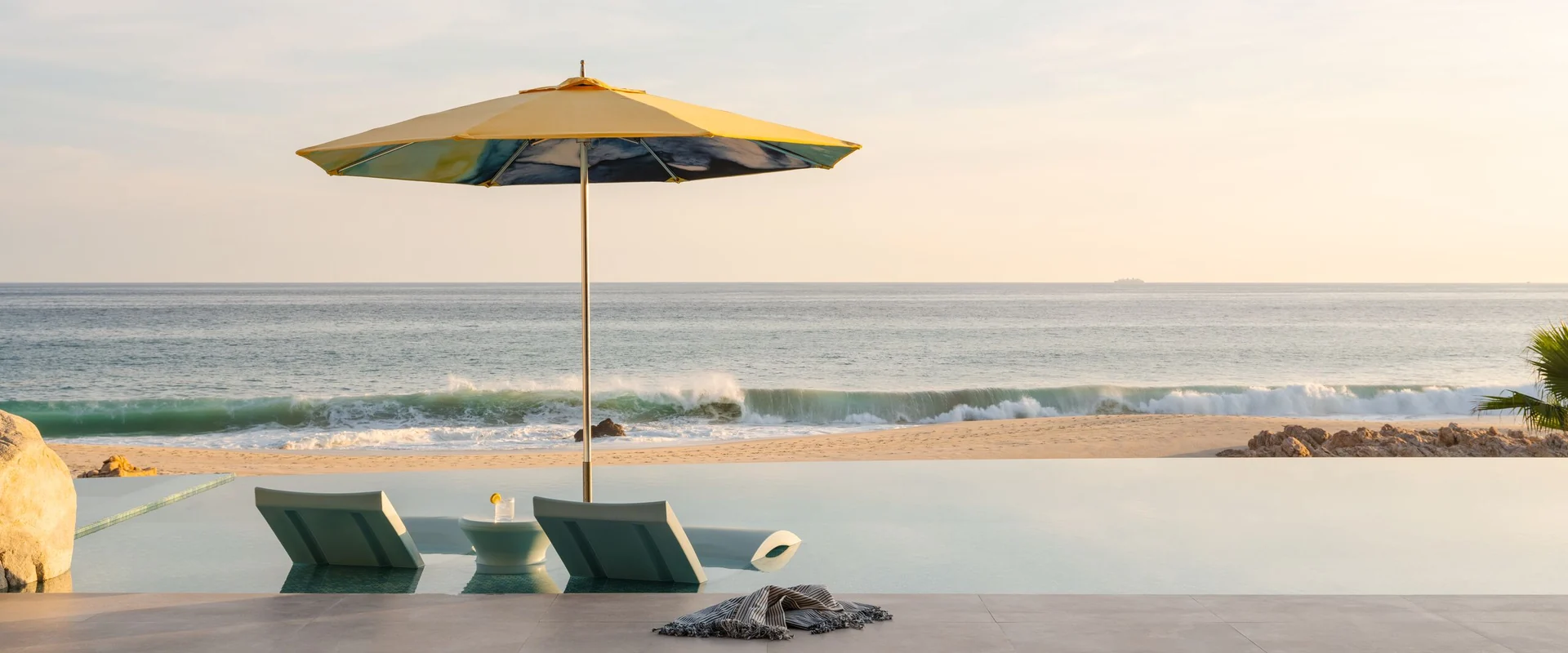 Yellow Tuuci parasol shading two in-pool loungers at the edge of an infinity pool, with waves breaking on the beach beyond at sunset
