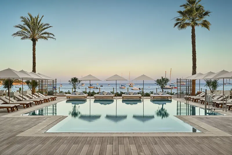 Rows of Tuuci Bay Master M1 Fiber-Flex parasols over sun loungers around a swimming pool on a wooden deck, with palm trees and boats moored in the bay behind, in Ibiza