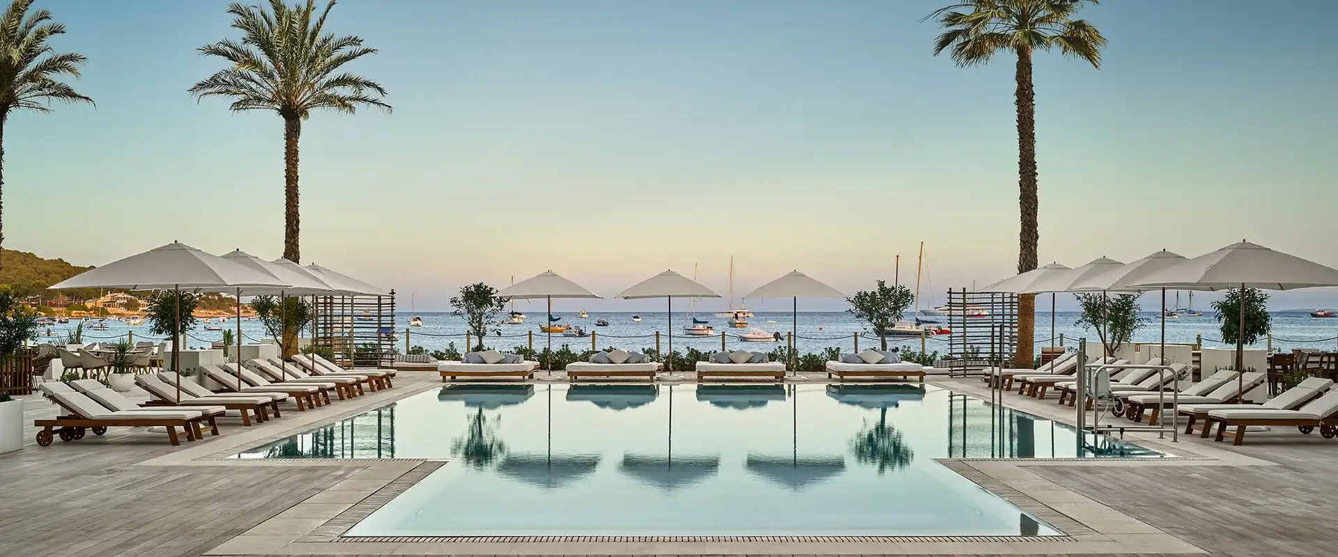 Rows of Tuuci Bay Master M1 Fiber-Flex parasols over sun loungers around a swimming pool on a wooden deck, with palm trees and boats moored in the bay behind, in Ibiza