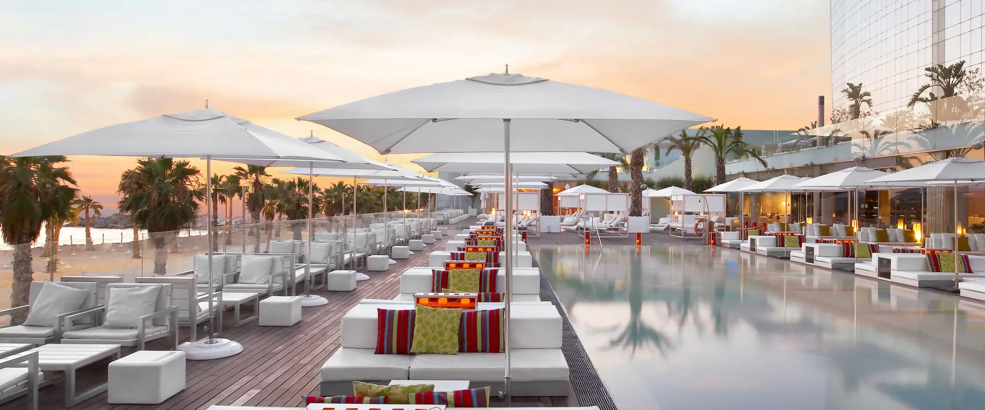 Rows of white Tuuci Ocean Master M1 parasols over a seafront hotel pool terrace in Barcelona at sunset