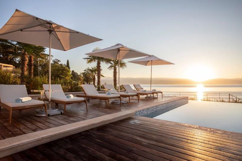 Square Tuuci parasols over teak sun loungers on a deck beside an infinity pool at sunset, with the sea and palms behind
