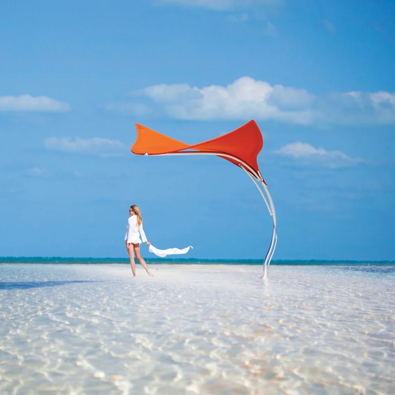 Red Tuuci Stingray shade sculpture on a curved polished mast standing in clear shallow water over white sand, with a woman in white walking beside it under a blue sky