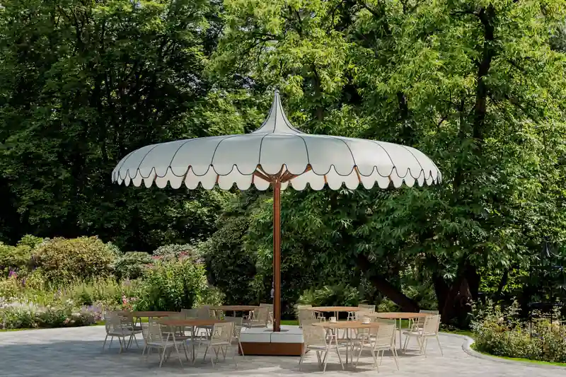White Tuuci parasol with a scalloped VOILARE canopy and teak pole shading a table and chairs on a stone terrace in a leafy garden