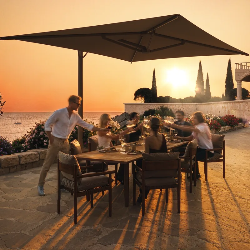 Dark square Umbrosa Aurelis cantilever parasol above friends dining on a stone terrace by the sea at sunset