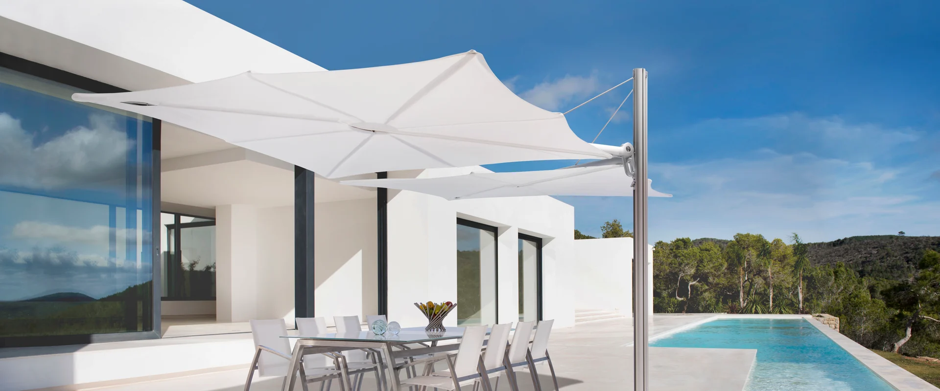 White Umbrosa Duo Spectra cantilever parasol shading an outdoor dining table on the terrace of a white modern villa, beside a long pool with pine-covered hills beyond