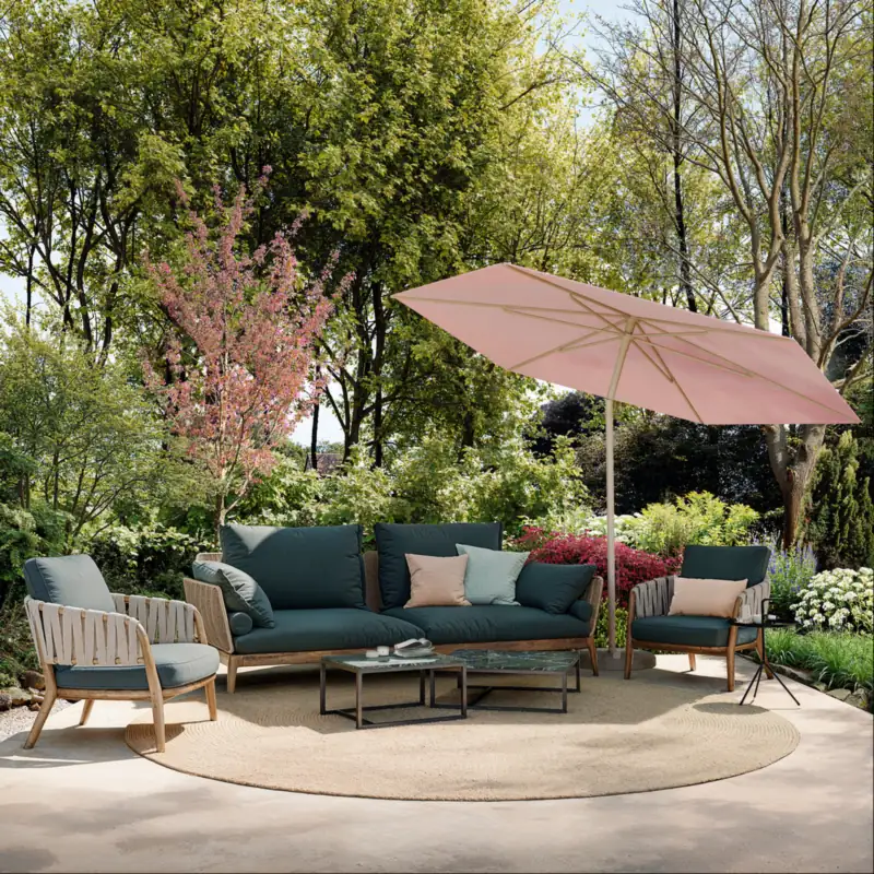 Tilted blush-pink Umbrosa Nata center-pole parasol shading a teal sofa and armchairs on a round rug in a leafy garden