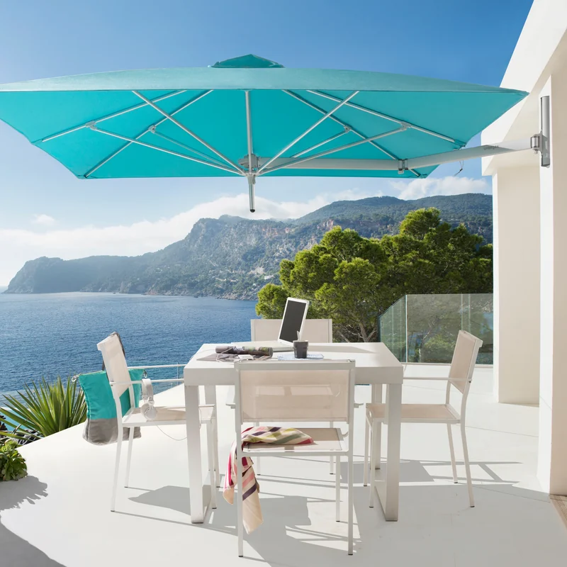 Turquoise Umbrosa Paraflex wall-mounted parasol over a white dining table on a terrace overlooking the sea and cliffs