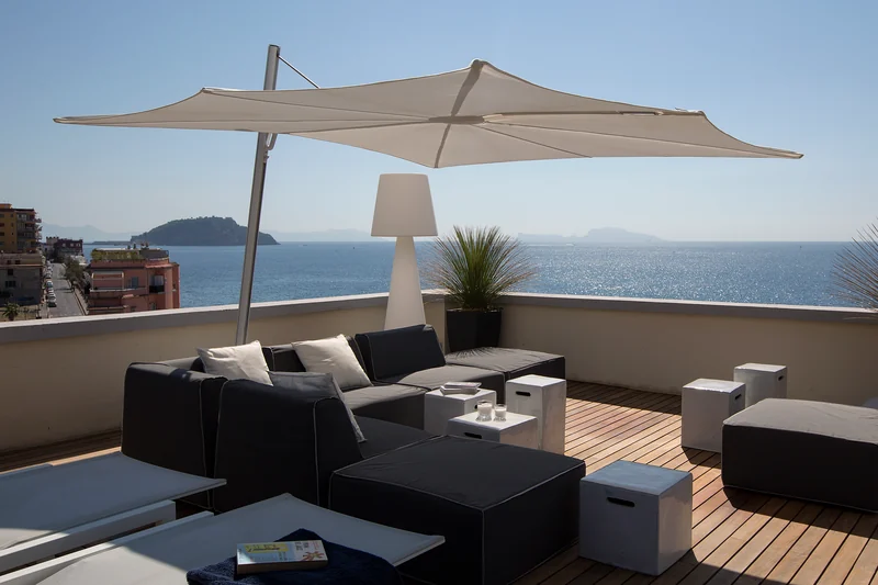 White Umbrosa Spectra cantilever parasol over dark lounge sofas on a rooftop terrace, with the sea and islands on the horizon