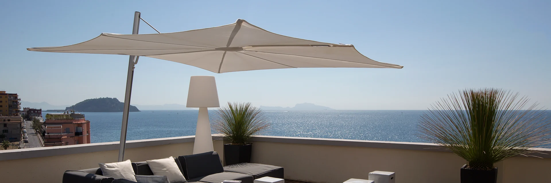 White Umbrosa Spectra cantilever parasol over dark lounge sofas on a rooftop terrace, with the sea and islands on the horizon