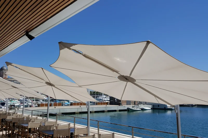 Row of white Umbrosa Spectra cantilever parasols seen from below on a waterfront terrace beside a marina, under a clear blue sky