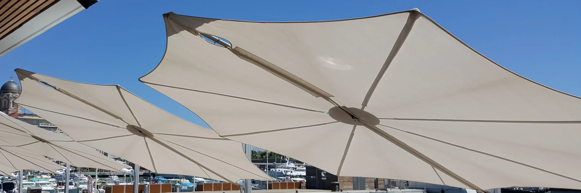 Row of white Umbrosa Spectra cantilever parasols seen from below on a waterfront terrace beside a marina, under a clear blue sky