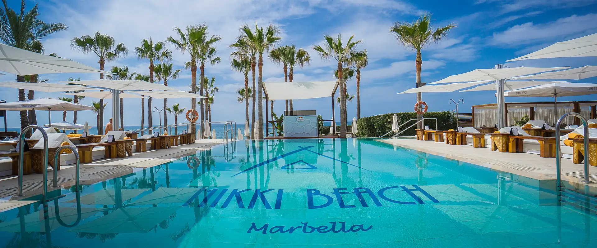 White Umbrosa Spectra cantilever parasols shading daybeds beside the pool at Nikki Beach Marbella, with palm trees and the sea behind