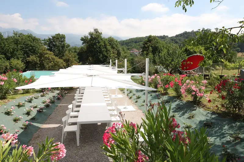 Row of white Umbrosa Spectra parasols over a long white table in a garden with oleanders, a pool and wooded hills behind