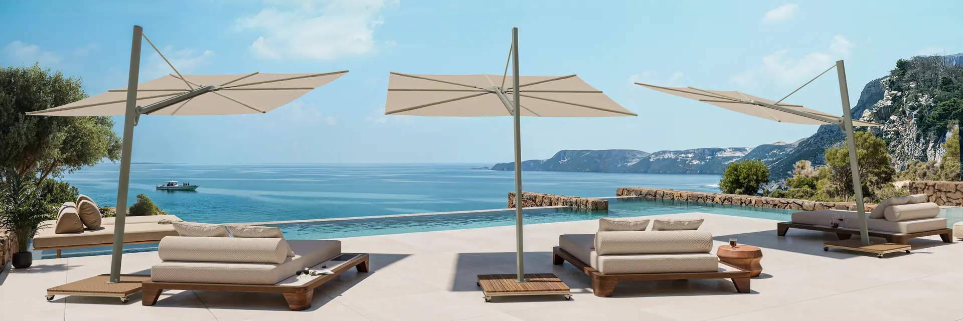 Sand-coloured Umbrosa Versa cantilever parasols shading daybeds on a stone terrace above the sea, with cliffs along the coast