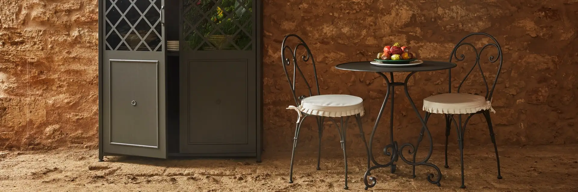 Unopiù Aurora wrought-iron table and chairs in front of an old brick wall with an iron-framed door and a lantern